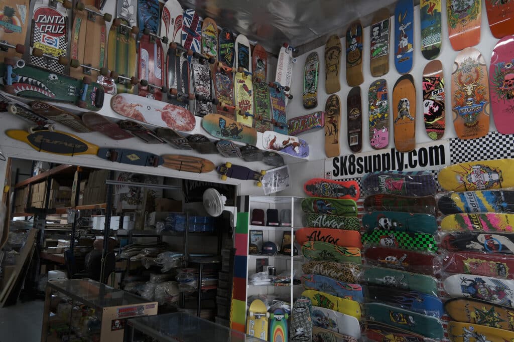 Shapers-Club- A diverse collection of colorful skateboards and surfboards displayed on a wall with "sk8supply.com" signage. -surfshop-surfboard