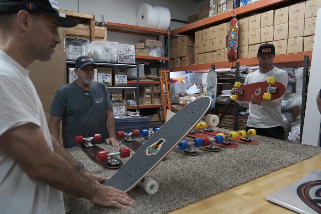 Shapers-Club- Men examining a Gordon Smith skateboard in a warehouse. -surfshop-surfboard