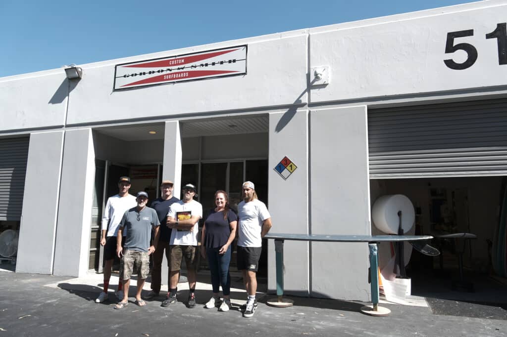 Shapers-Club- Group of individuals posing together in front of an industrial building with a sign reading "Gordon and Smith Custom Surf. -surfshop-surfboard