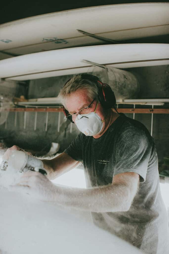Shapers-Club- Roger Hinds, wearing a dust mask and hearing protection, shapes a surfboard in a workshop, with dust particles visible in the air. -surfshop-surfboard