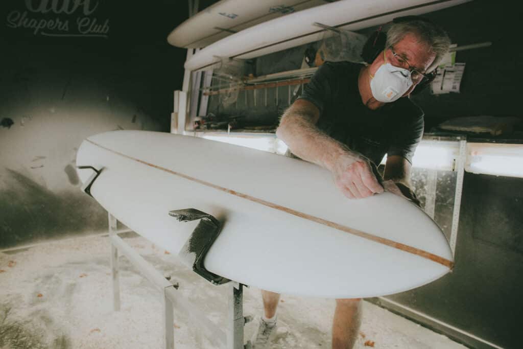 Shapers-Club- Roger Hinds, wearing a dust mask and goggles, shapes a surfboard in the Shape Universe workshop with surfboards in the background. Dust particles are visible in the air. -surfshop-surfboard
