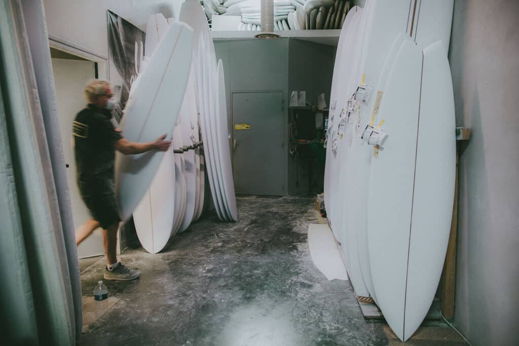 Shapers-Club- A person walks through a workshop, the Universe of Shape, where several surfboards made by Master Surfboard Craftsman Roger Hinds are lined up upright against the wall. -surfshop-surfboard
