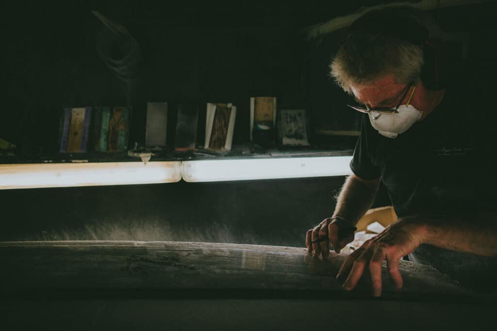 Shapers-Club- A person wearing protective gear works meticulously on a piece of fabric at Univers du Shape, in a dimly lit workshop. -surfshop-surfboard