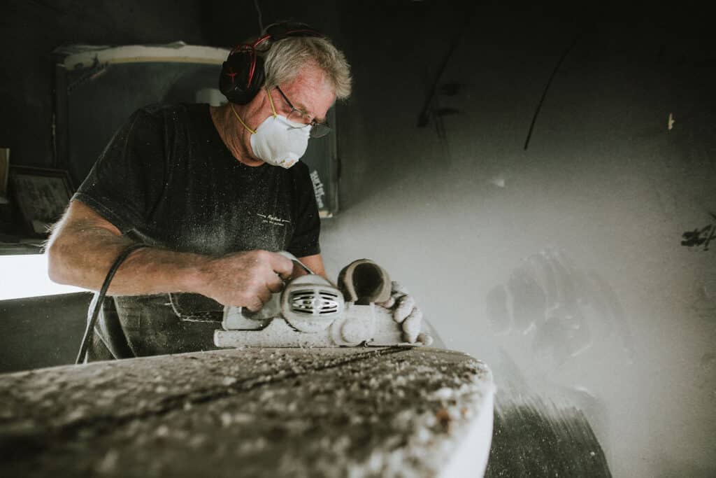 Shapers-Club- A person wearing a dust mask and goggles uses a circular saw, cutting a slab of stone for the Shape Universe and generating a significant amount of dust. -surfshop-surfboard