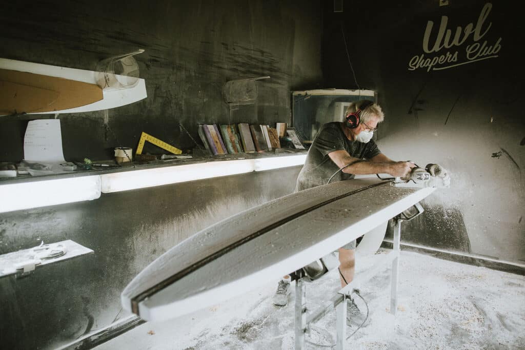 Shapers-Club- A Master Surfboard Craftsman wearing protective gear shapes a surfboard in a dusty workshop. -surfshop-surfboard