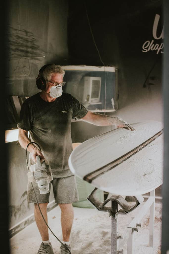 Shapers-Club- A person wearing protective gear uses a power tool on Roger Hinds surfboards in a workshop, with dust particles visible in the air. -surfshop-surfboard