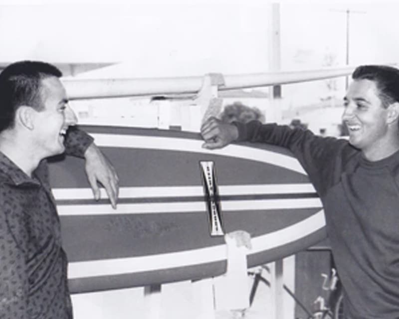 Shapers-Club- Two smiling men leaning on a Gordon & Smith surfboard. -surfshop-surfboard
