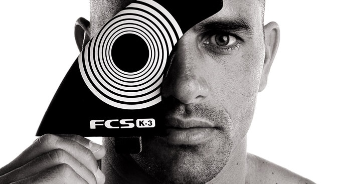 Shapers-Club- Black and white photo of a man holding a card with concentric circles over one eye and staring at the camera. The card reads "fcs k-3. -surfshop-surfboard