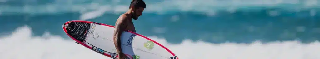 Shapers-Club- Un surfeur portant une planche avec le meilleur LEASH marchant le long du rivage avec des vagues en arrière-plan. -surfshop-surfboard