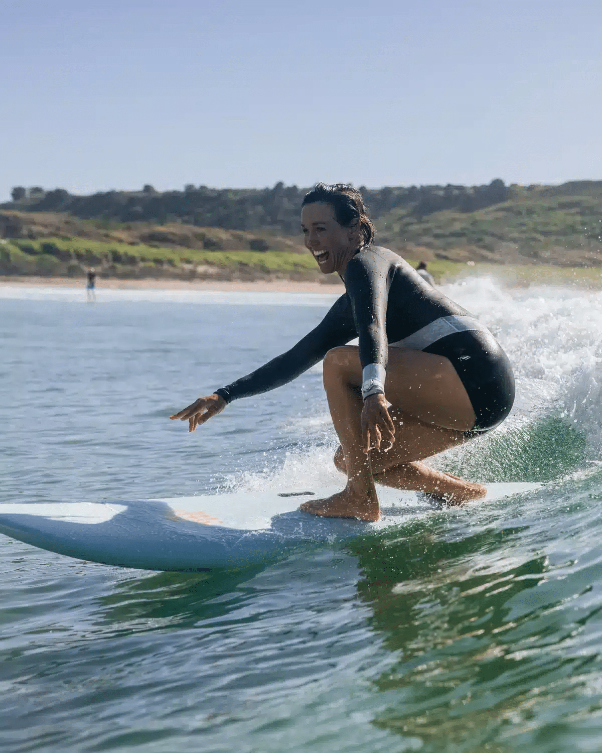 Shapers-Club- A wet-haired person wearing a black wetsuit rides a wave on a SOFTECH FB 7'0 surfboard, Handshaped Sally Fitzgibbons Mist, with the shoreline and distant hills visible in the background on a sunny day. -surfshop-surfboard