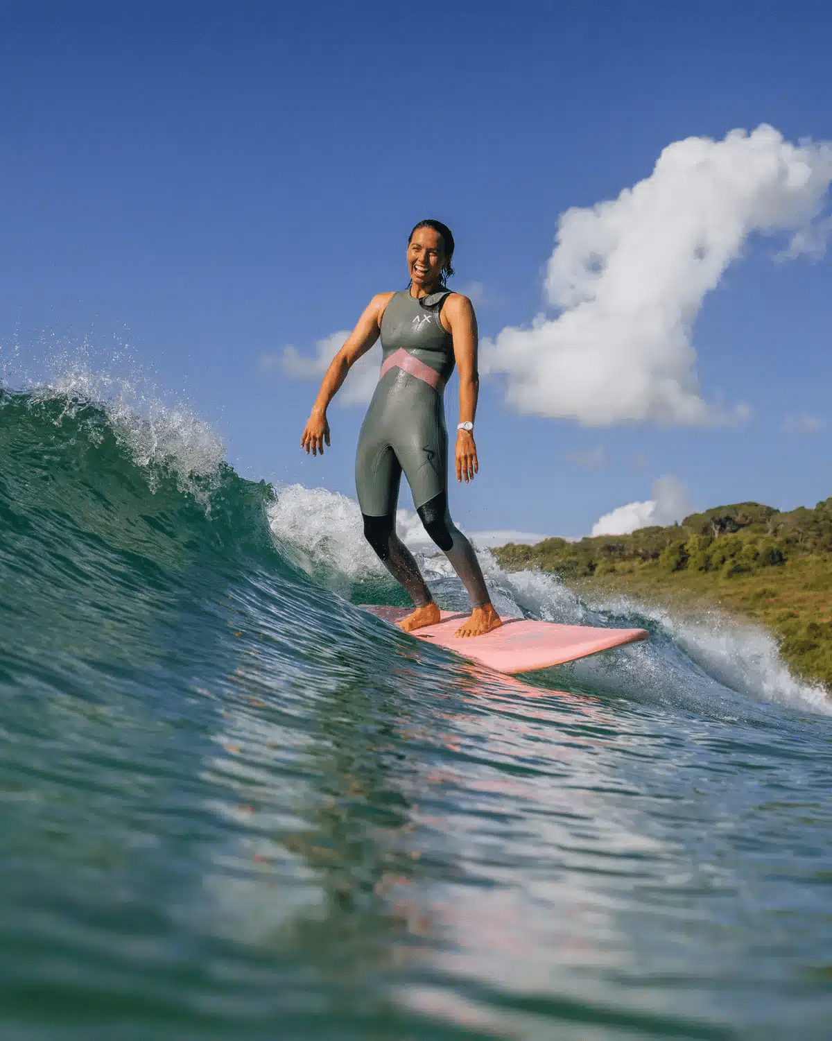 Shapers-Club- A person wearing a wetsuit surfs a wave with the Sally Fitzgibbons FB 7'0 Pink surfboard from SOFTECH - Hand shaped. The weather is clear and the sky is blue with a few clouds. There's a hill with greenery in the background. The person seems to be smiling and enjoying the ride. -surfshop-surfboard