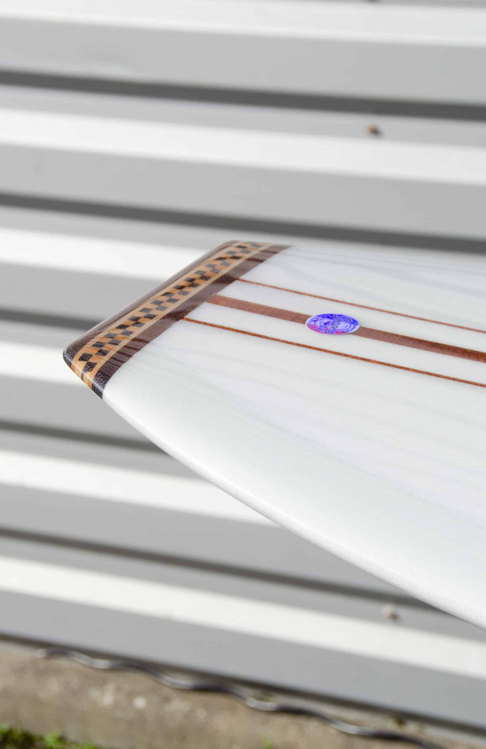 Shapers-Club- Close-up of the tail of Roger Hinds' "The Classic" surfboard, measuring 9'4" x 23" x 3' 1/4", with brown and black stripes and a small circular blue sticker. The background features gray and white horizontal stripes, probably on a metal or wood wall. -surfshop-surfboard