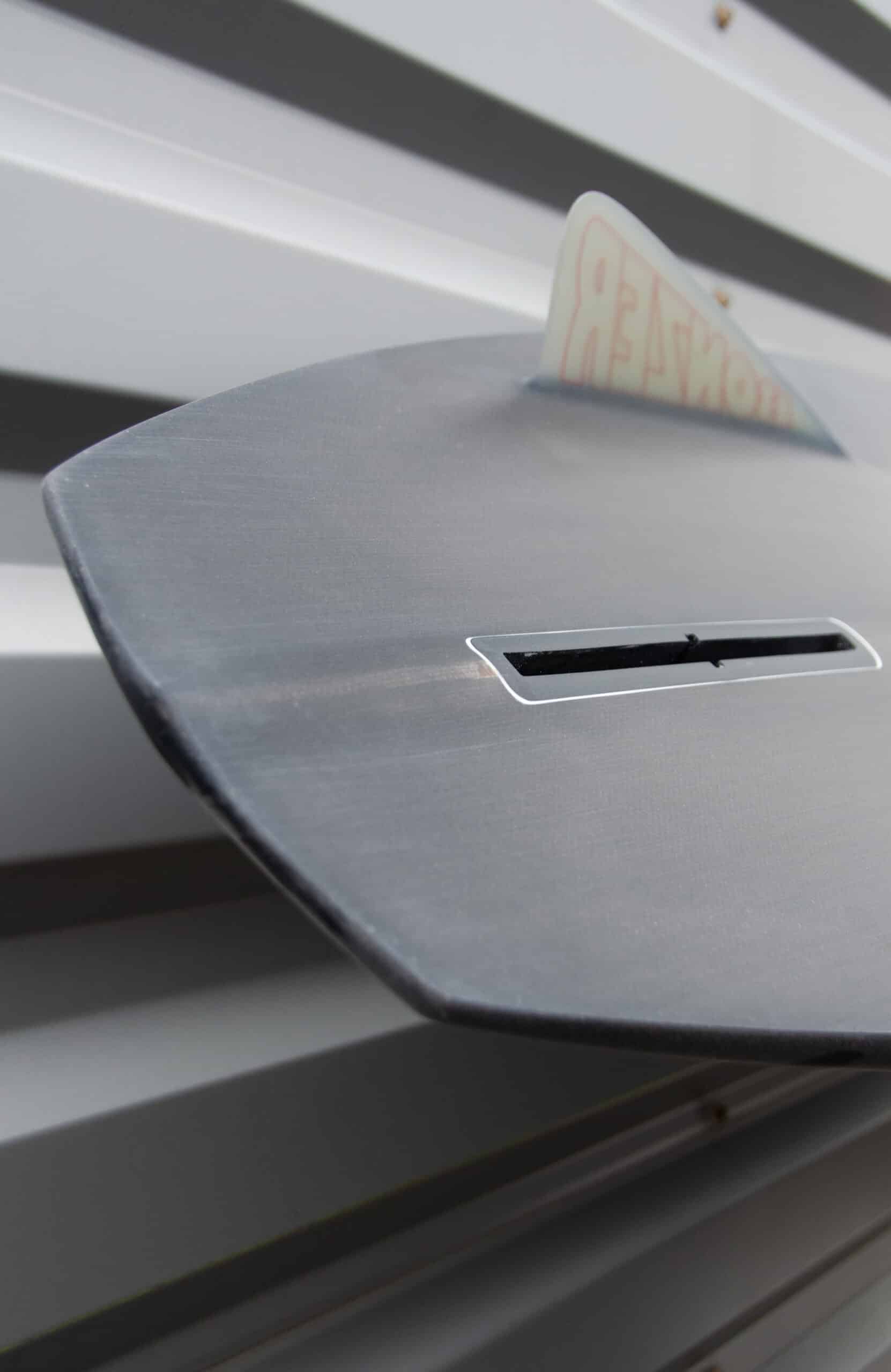 Shapers-Club- Close-up of a gray envelope mailbox with partial view of a partially inserted white envelope. The mailbox, whose design reflects the clean lines characteristic of the Campbell Brothers surfboard - Russ Short 7'0" x 20' 3/4" x 2' 3/4", is mounted on a horizontally ribbed metal surface. -surfshop-surfboard
