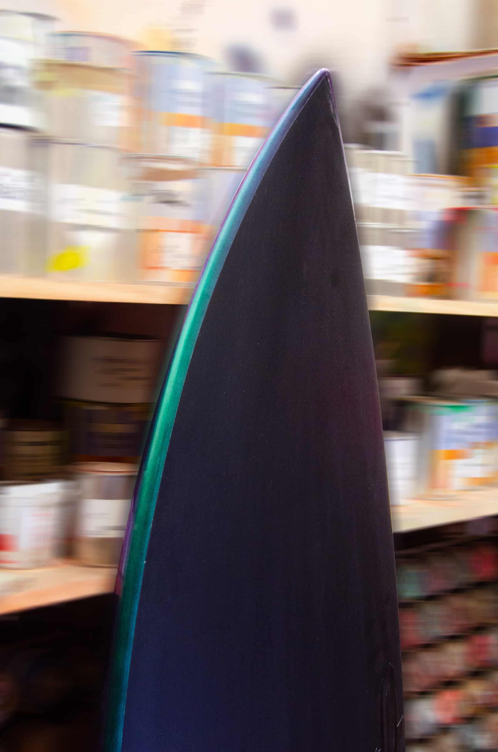 Shapers-Club- A close-up of the tip of a Fire Arrow surfboard highlights its elegant design with a subtle color gradient. This 5'9" x 19' 1/4" x 2' 1/2" black board is designed for speed and agility on every wave, with shelves full of cans, jars and miscellaneous containers in the background. -surfshop-surfboard