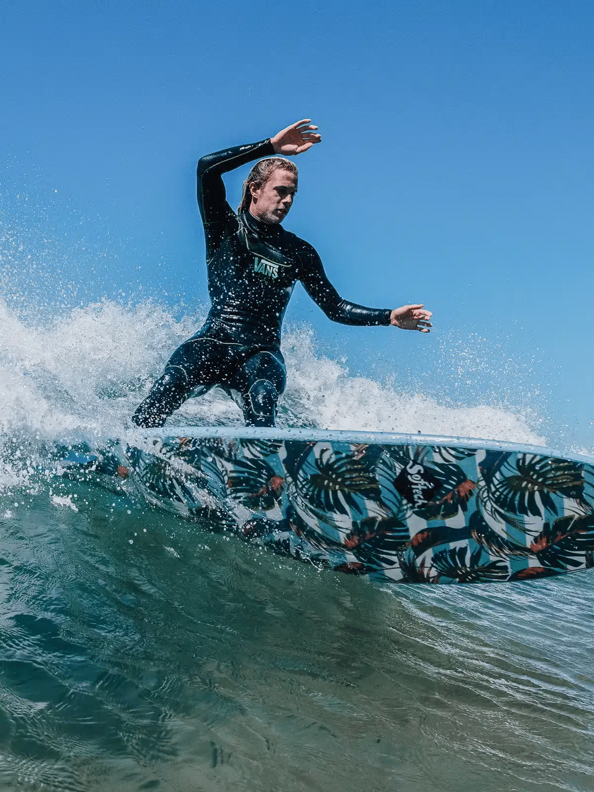 Shapers-Club- Sally Fitzgibbons, dressed in an elegant wetsuit, surfs a wave on her handcrafted Sally Fitzgibbons FB 7'0 Mist surfboard with a tropical leaf motif. The sky is clear and blue, and the water splashes around her as she deftly maneuvers on the wave. -surfshop-surfboard