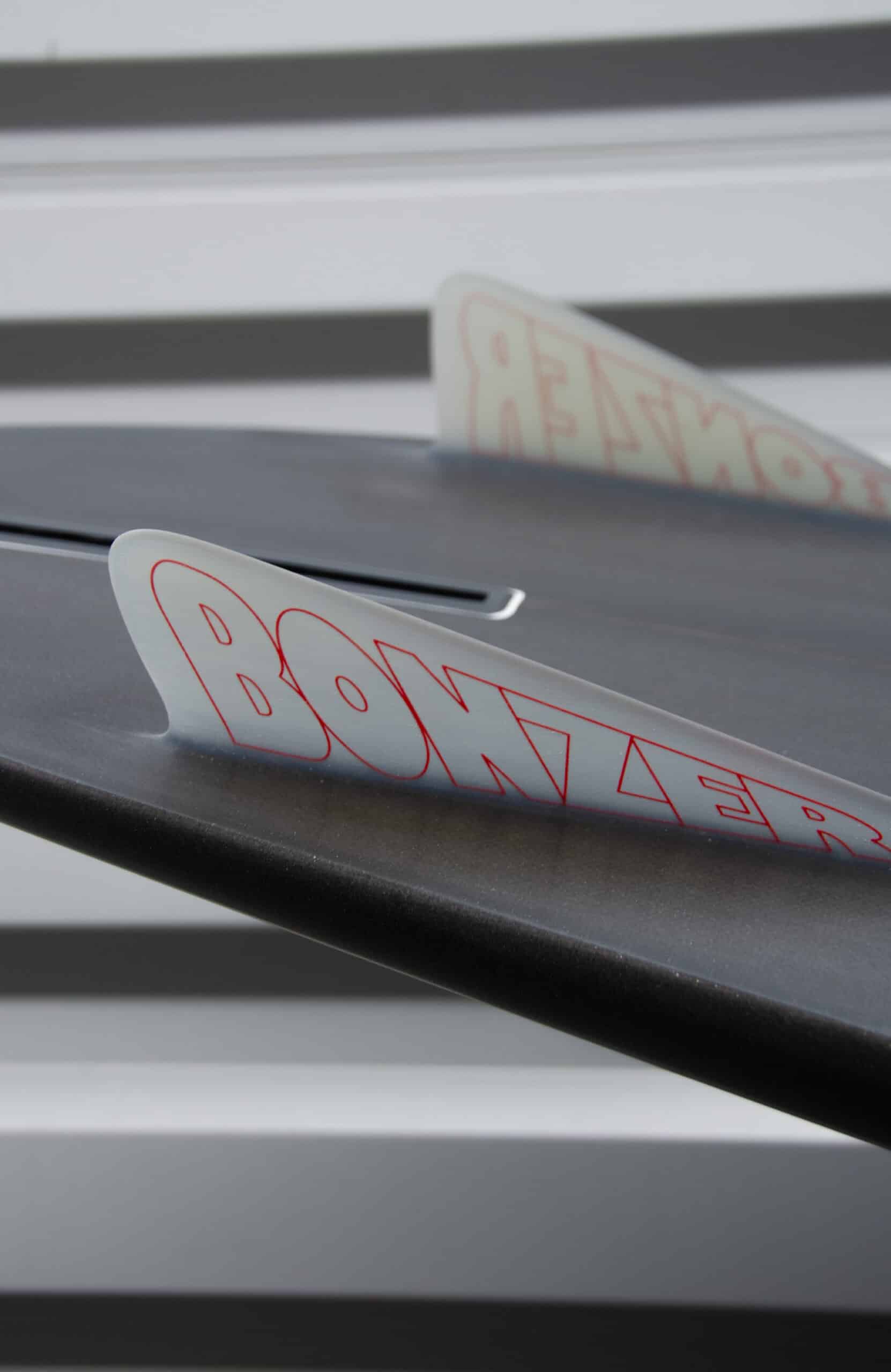 Shapers-Club- Close-up of a Campbell Brothers - Russ Short surfboard fin with the word "Bonzer" written on it in red letters. The background features blurred gray and white horizontal stripes. The focus is on the fin, offering a partial view of the elegant design of the 7'0" x 20' 3/4" x 2' 3/4" surfboard. -surfshop-surfboard