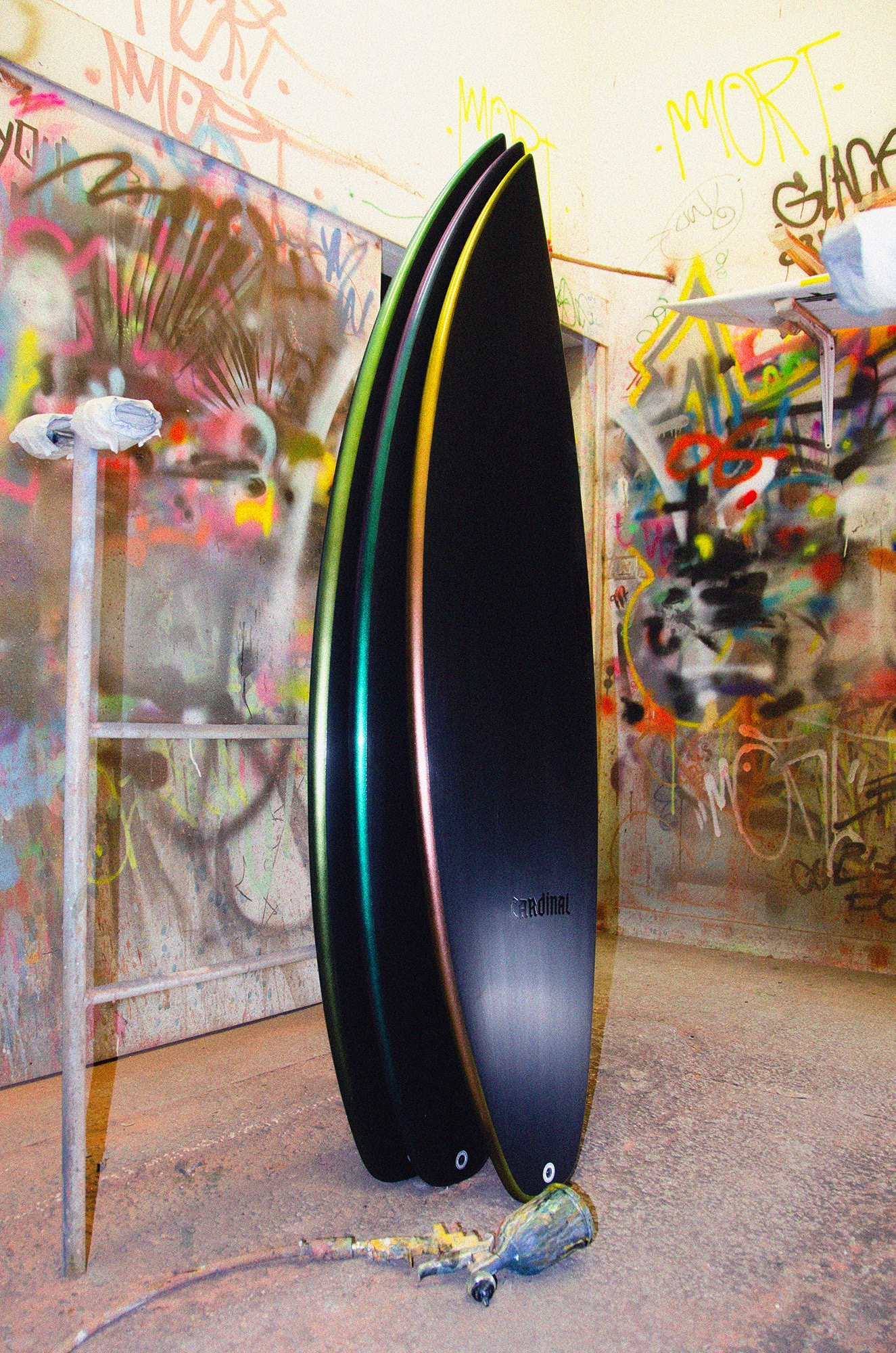 Shapers-Club- In a graffiti-covered room, two black Fire Arrow surfboards measuring 6'0" x 20" x 2' 5/8" with colored edges stand upright. Nearby, a metal stand and a few painting tools are scattered on the floor. -surfshop-surfboard