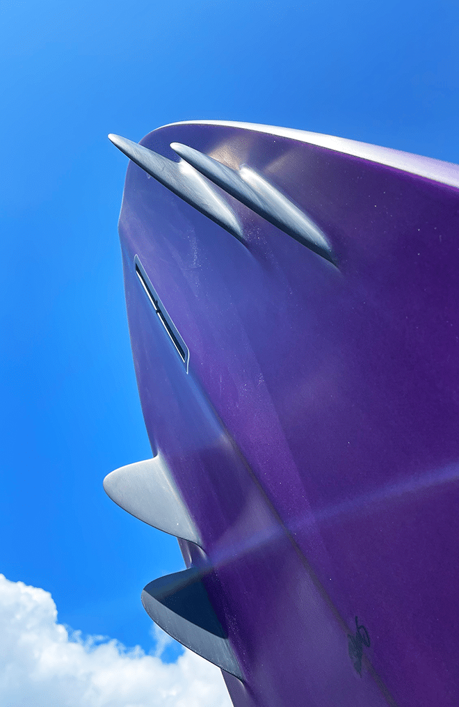 Shapers-Club- Close-up of the Mini Bonzer surfboard 5'8" x 21' 1/4" x 2' 11/16" against a bright blue sky, highlighting its elegant fins and small logo. A few clouds are visible in the lower corner. -surfshop-surfboard