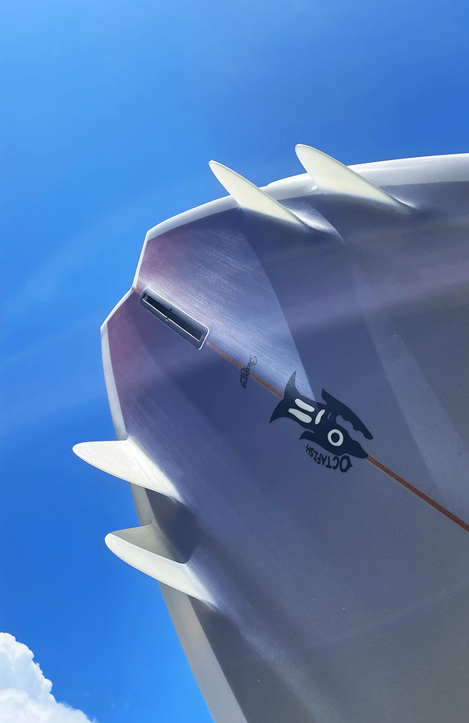 Shapers-Club- A 5'10" Octafish surfboard, featuring a sleek white surface with multiple fins, is photographed from below against a bright blue sky background. It features a striking black graphic design and bears the "02" logo as well as the name "Octafish", as it glides beneath fluffy clouds at the edge of the image. -surfshop-surfboard