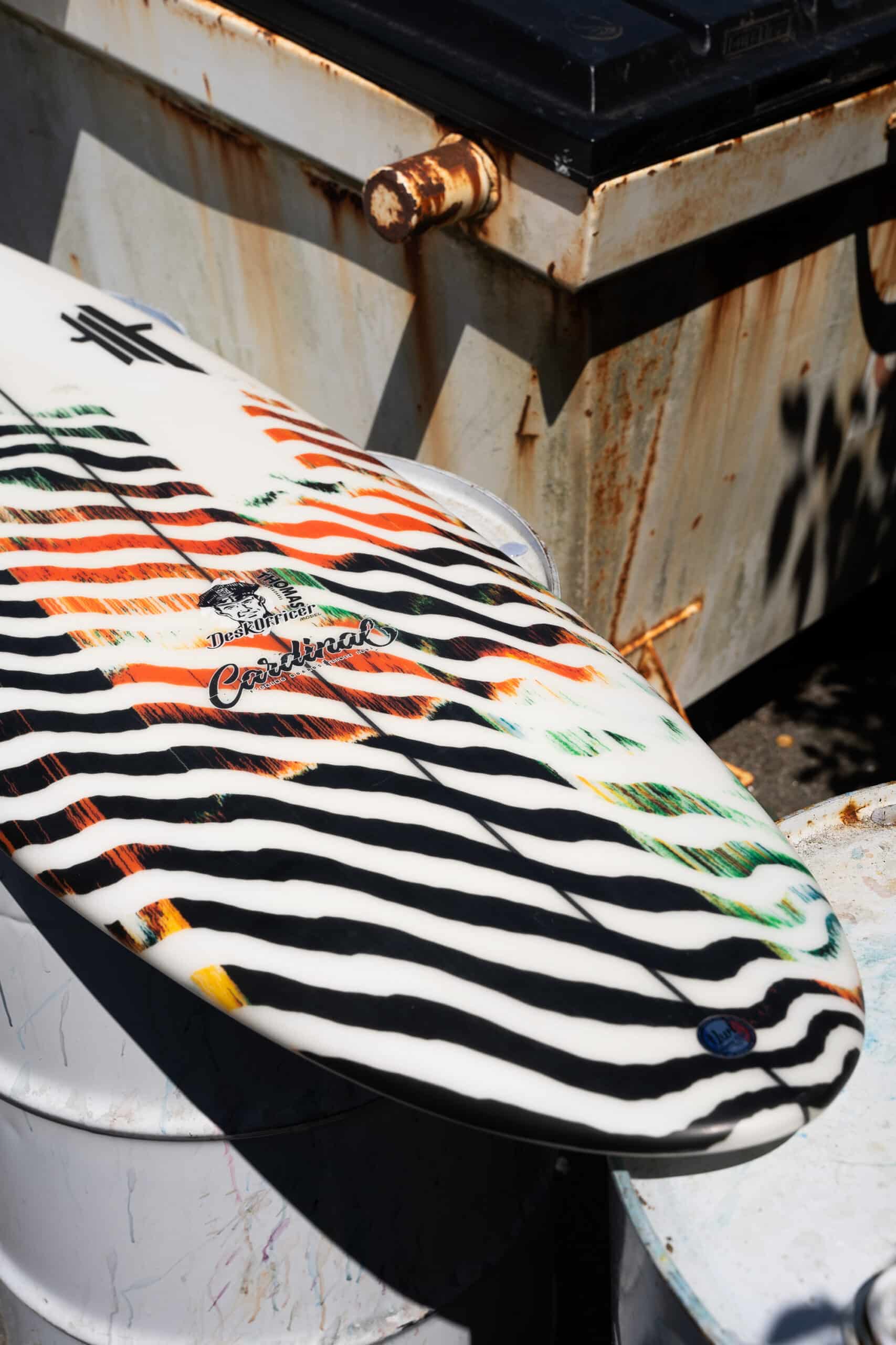 Shapers-Club- La planche de surf Desk Officer, 5'7" x 19" x 2' 3/8", arbore des rayures zébrées noires et blanches audacieuses avec des touches d'orange et de vert, appuyées contre des barils en métal rouillé. Elle mélange parfaitement les ambiances industrielles et sportives, créant une scène dont même les officiers de bureau d'intérieur pourraient rêver au milieu de la paperasse. -surfshop-surfboard