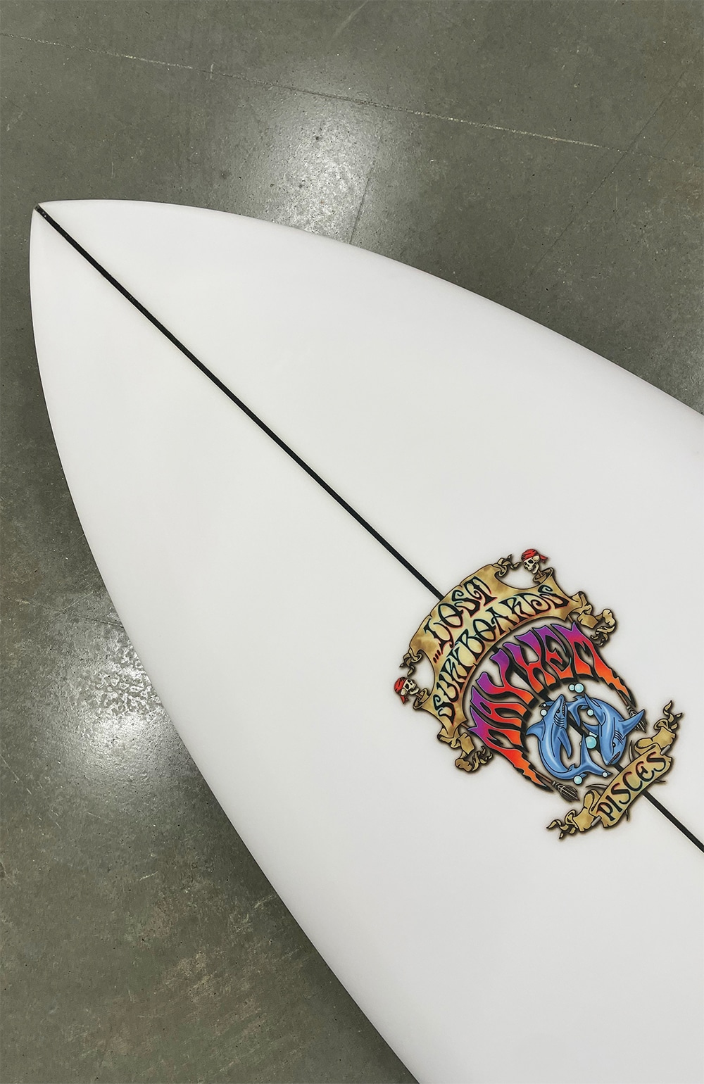 Shapers-Club- A close-up of the white Pisces surfboard highlights a dynamic design near the tip, adorned with detailed illustrations and phrases such as "Lost" and "Psycho Killer". This board, with its 38.2 L volume, is elegantly displayed on a polished concrete floor. -surfshop-surfboard