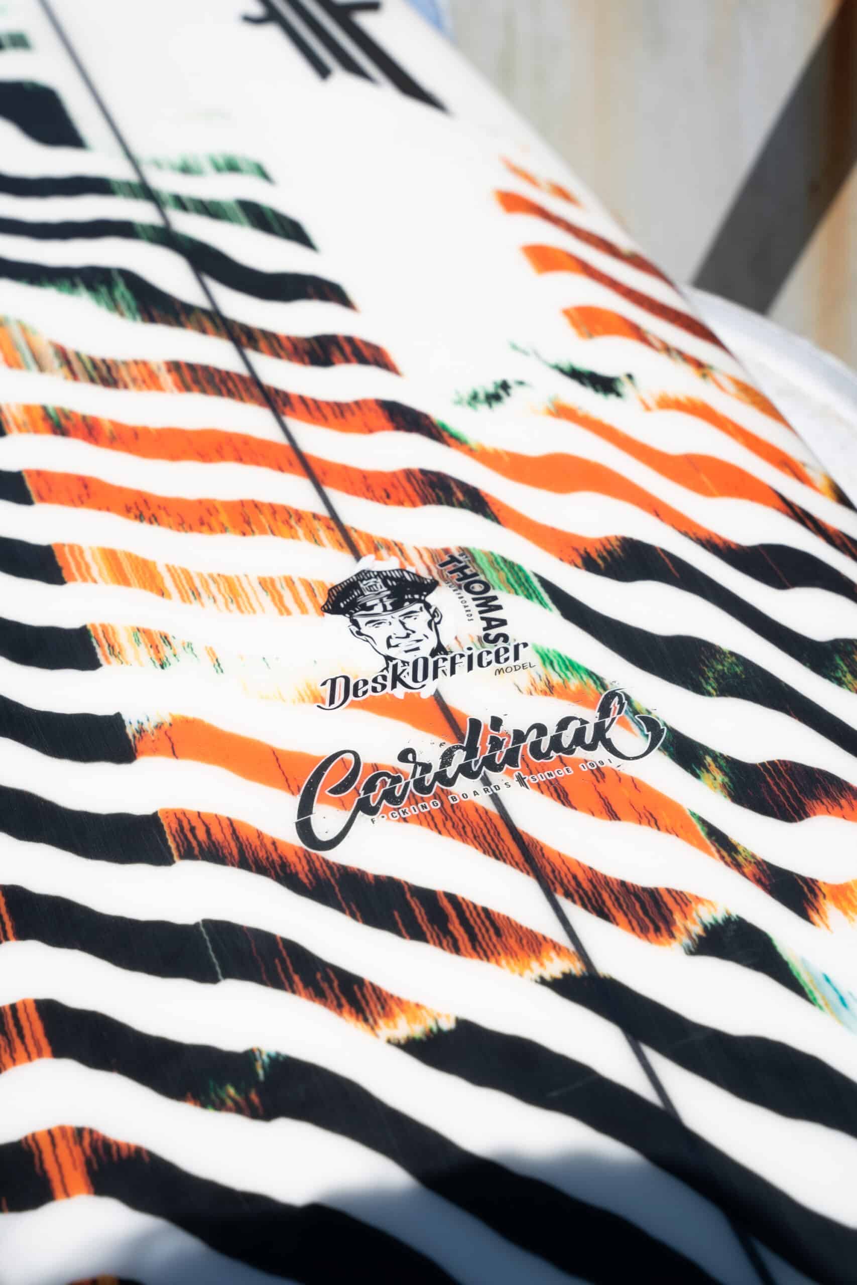 Shapers-Club- Gros plan d'une planche de surf aux rayures orange, vertes et noires éclatantes. Elle arbore les logos « The Mason Desk Officer » et « Caribbean », avec des dimensions élégantes de 5'7" x 19" x 2' 3/8", ce qui renforce son attrait dynamique. -surfshop-surfboard