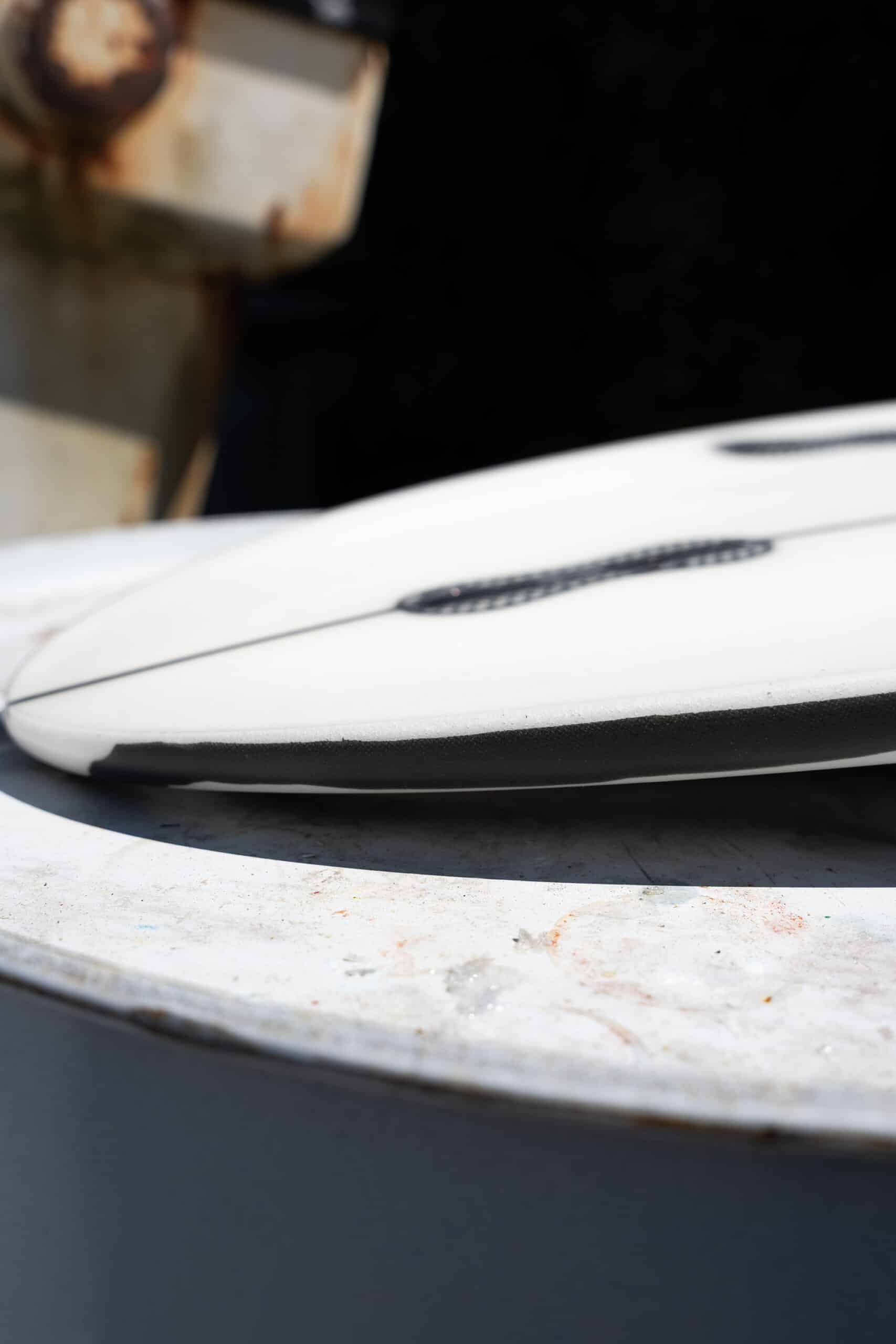 Shapers-Club- Gros plan sur la surface blanche et noire de la planche de surf Desk Officer, posée sur du béton, faisant écho aux lignes du mobilier de bureau, avec un arrière-plan en métal rouillé. Ses dimensions de 5'7" x 19" x 2'3/8" créent des rainures visibles uniques. -surfshop-surfboard