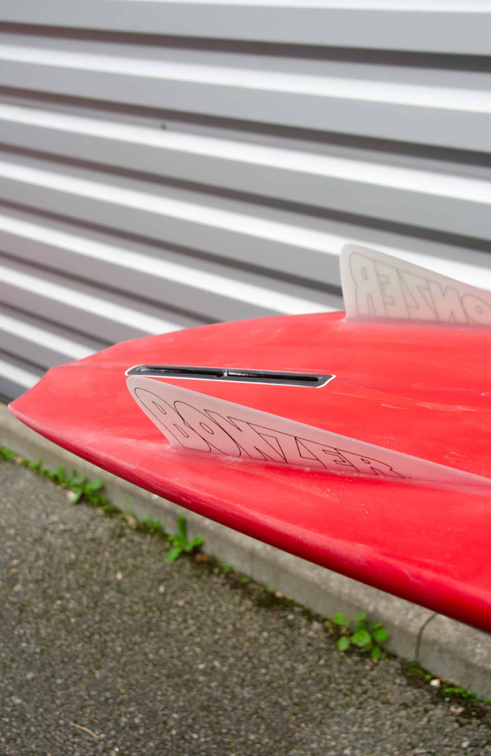 Shapers-Club- Close-up of Russ Short surfboard tail in red with fin and "BONZER" displayed, measuring 6'2" x 19' 1/2" x 2' 1/2", on gravel near a corrugated iron wall. -surfshop-surfboard